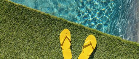 A pair of yellow flip-flops on a green artificial grass surface next to a clear blue pool.