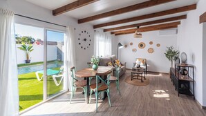 A spacious living area with a wooden floor, a dining table with chairs, a sofa, and a coffee table. There is a large window with curtains, a clock on the wall, and a small garden visible outside. - Casa Flamenquita Nerja by Ruralidays (Málaga)