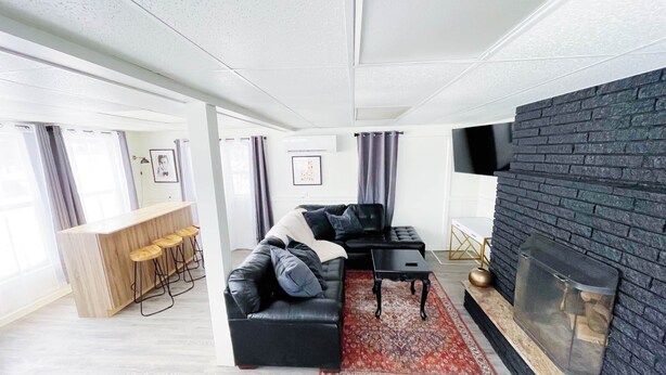 A modern living room with a black leather sofa, a coffee table, and a fireplace. There is a bar area with stools and a large window with curtains. - The Club House - Suite 101 (Sault Ste. Marie)