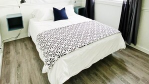 A bedroom with a bed, a wall-mounted abstract artwork, a window with curtains, and a bedside table with a lamp. - The Club House - Suite 101 (Sault Ste. Marie)