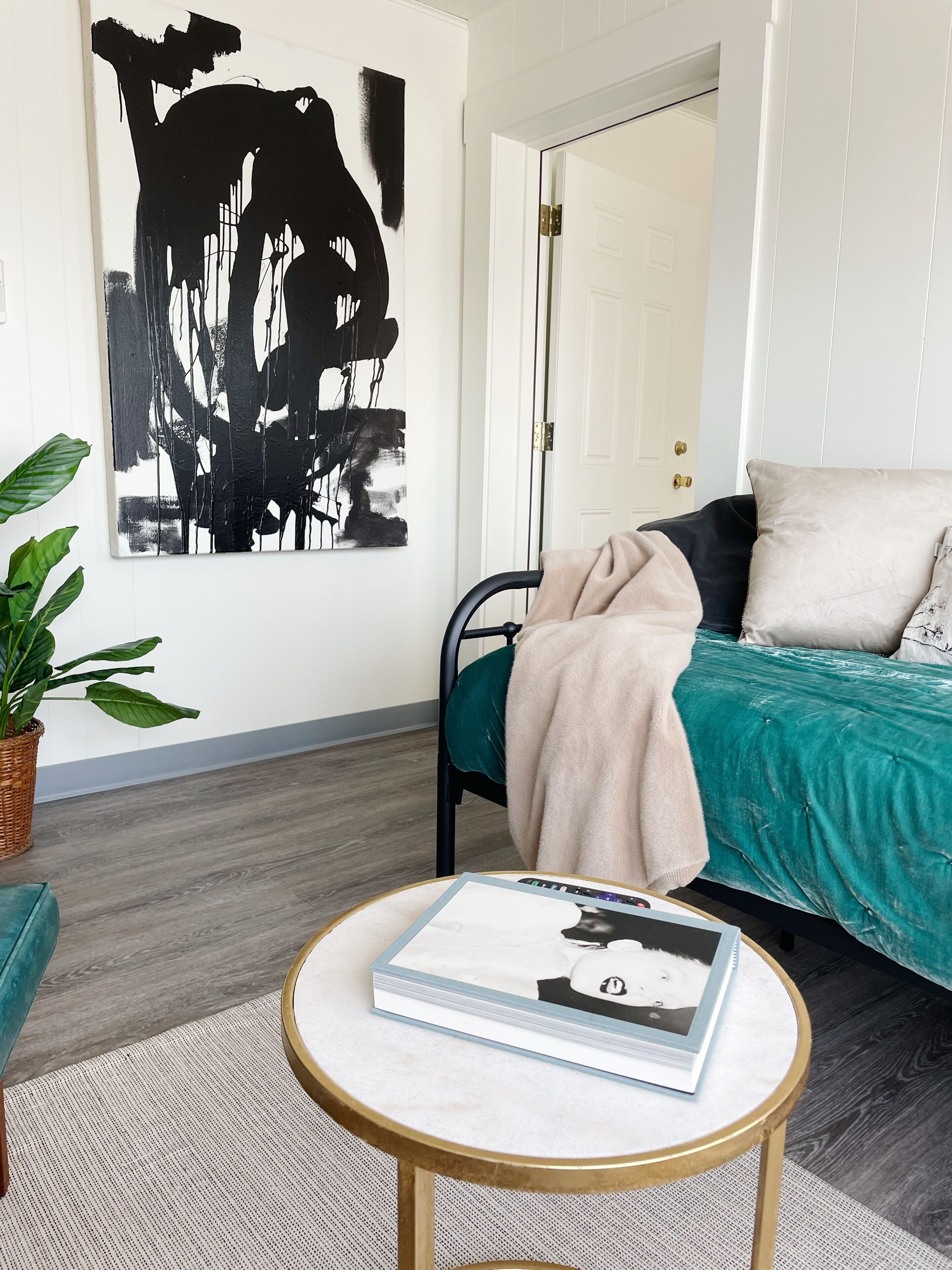 Een moderne slaapkamer met een blauwgroene sprei, een groot abstract schilderij aan de muur, een ronde salontafel met een boek, en een kamerplant.