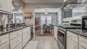 Una cocina moderna con armarios blancos, encimera de granito y electrodomésticos de acero inoxidable. Cuenta con un comedor con una mesa y sillas de madera.