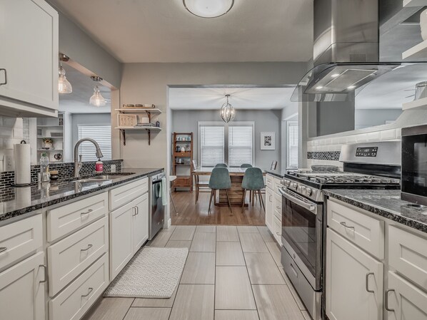 A modern kitchen with white cabinetry, a granite countertop, and stainless steel appliances. There is a dining area with a wooden table and chairs. - Charming home near Fairgrounds & DownTown sleeps 9 (Oklahoma City)