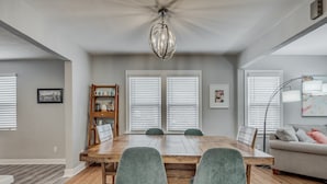 Comedor con mesa de madera y dos sillas verdes, junto a ventanas con persianas.