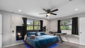 Un dormitorio con cama, ventilador de techo, mesita de noche, silla y ventana con cortinas.