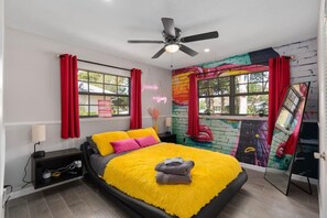 A bedroom with a bed, a ceiling fan, a window with red curtains, a wall mural, and a mirror.