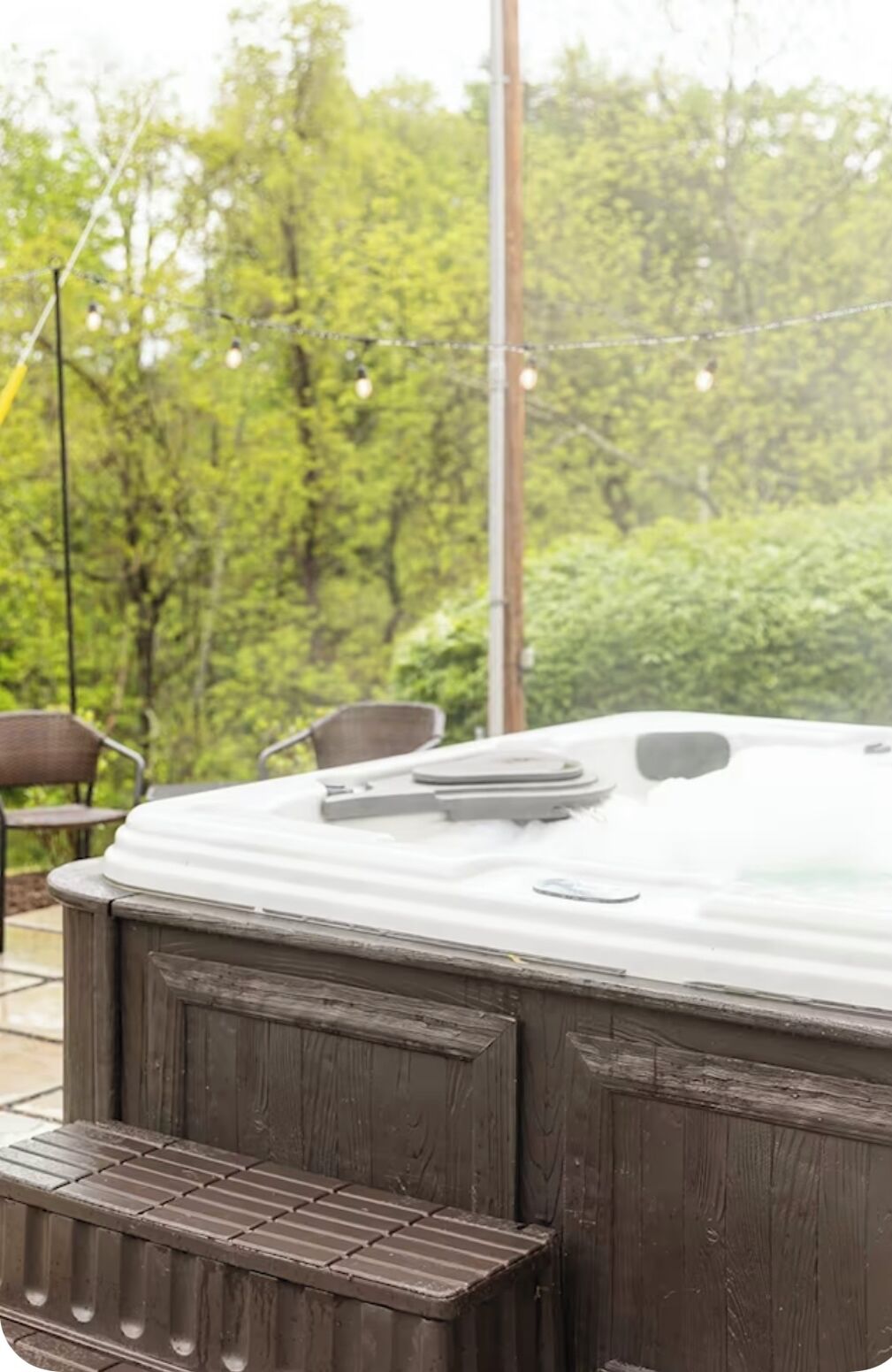 Un jacuzzi con revestimiento de madera, rodeado de vegetación y mobiliario de exterior.