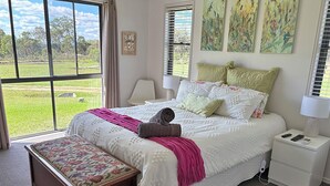 A bedroom with a large bed, a bedside table, a nightstand, a bench, and a window with a view of a green landscape. - Spacious Country Home (Mount Tully)