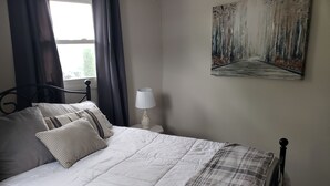 Un lit bien fait, avec une couette blanche et une couverture à carreaux, une lampe de chevet et un tableau accroché au mur.