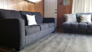 Une pièce avec deux canapés gris, dont l’un est agrémenté d’un coussin blanc et d’un mot écrit à la main, et une porte en bois sur laquelle est fixée une pancarte.