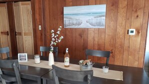 Un coin repas avec une table en bois, des chaises, un vase de fleurs, une bouteille, une bougie et un tableau encadré au mur.