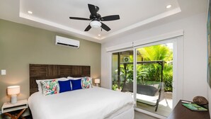 A bedroom with a bed, a ceiling fan, an air conditioning unit, a nightstand with a lamp, and a view of a balcony with outdoor furniture. - Surf, Relax and Explore the best of Avellanas (Playa Avellanas)