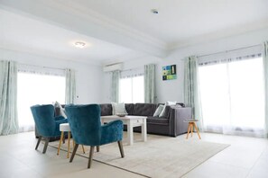 Un salon spacieux avec un canapé gris, deux fauteuils bleus, une table basse blanche et une petite table d’appoint.