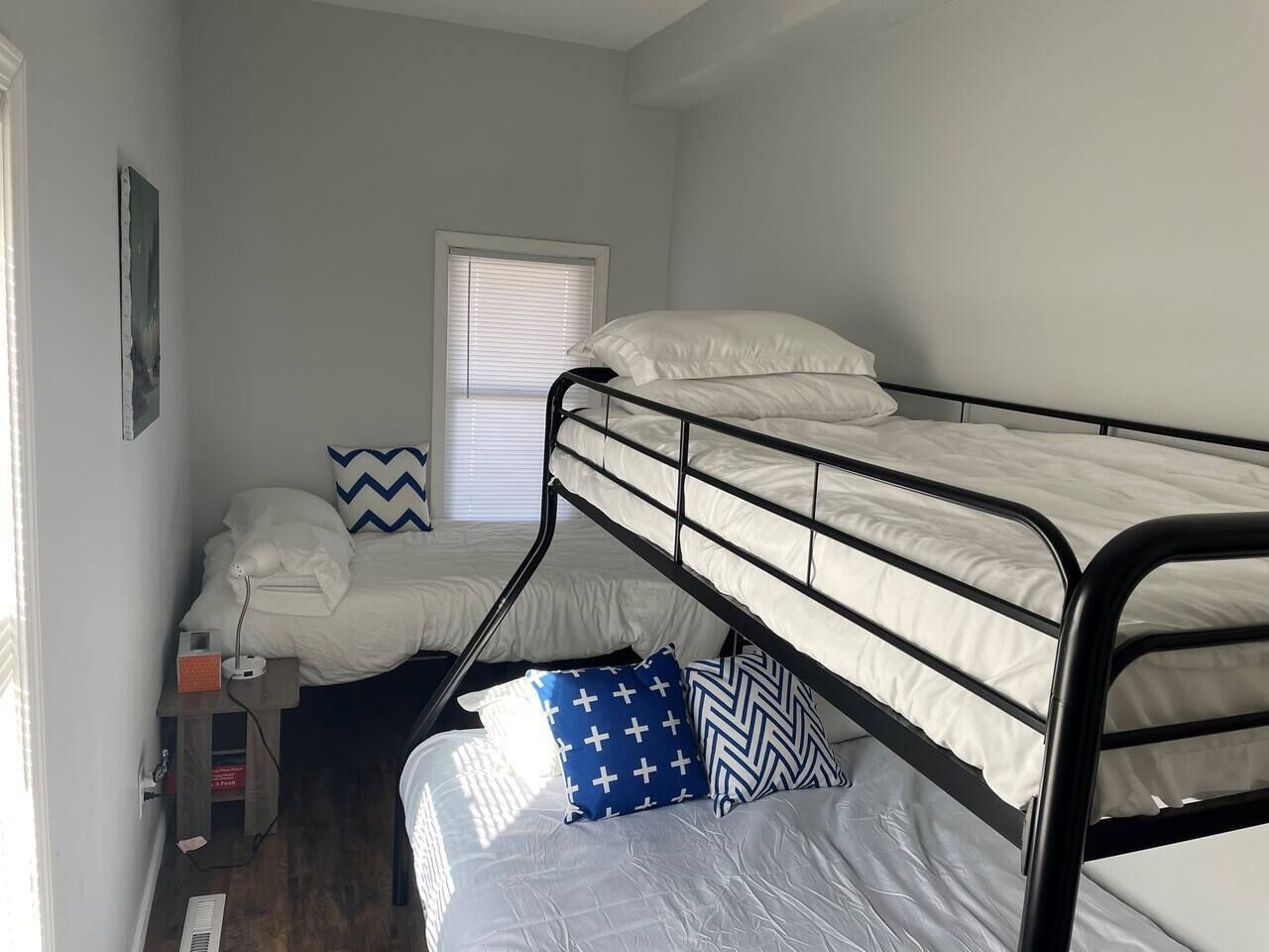 A dormitory room with bunk beds, a window with blinds, a small desk with a lamp, and a wall-mounted shelf.