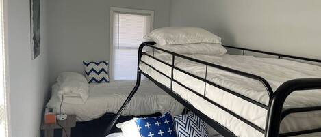 A dormitory room with bunk beds, a window with blinds, a small desk with a lamp, and a wall-mounted shelf.