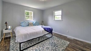 A bedroom with a bed, two windows, a nightstand, and a patterned rug. - Dunes Home – Walk to Town, Fenced Yard, Near Dunes | Valpo University | Dunes (Chesterton)