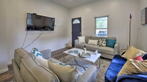 A living room with a flat-screen TV mounted on the wall, a sofa, a coffee table with books and a bottle, and a window with a view of the outside. - Dunes Home – Walk to Town, Fenced Yard, Near Dunes | Valpo University | Dunes (Chesterton)