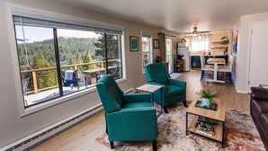 A spacious living area with large windows, a cozy seating area with teal chairs, a wooden coffee table, and a kitchen in the background. - Coeur d'Alene Lake House - Hot Tub with lake view, beach/lake access/boat launch (Worley)