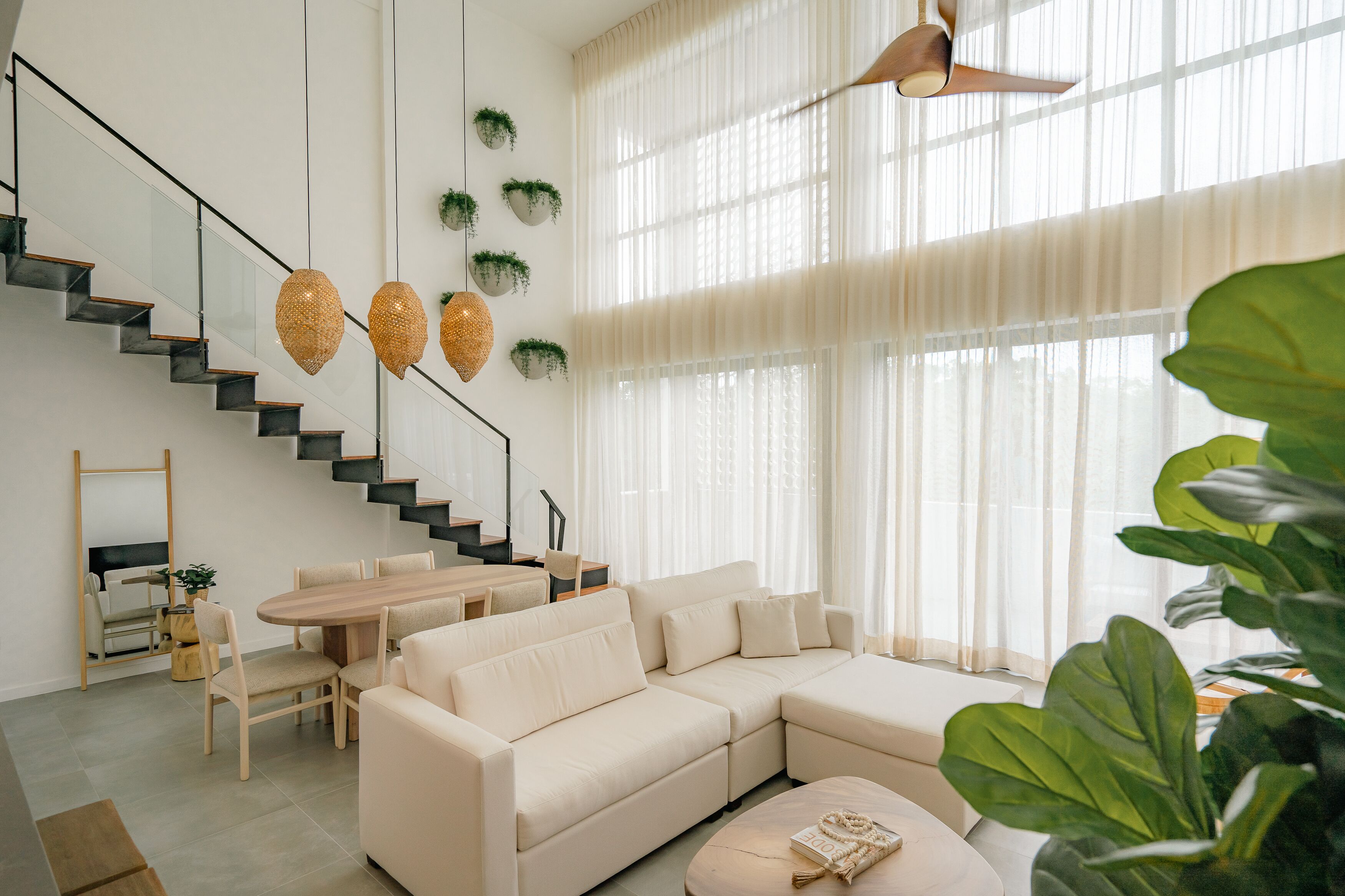 A modern living room with a large beige sectional sofa, a dining table with chairs, and a staircase with glass railings.
