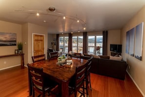 A spacious living area with a wooden dining table, comfortable seating, a television, and large windows offering a view of the outdoors.