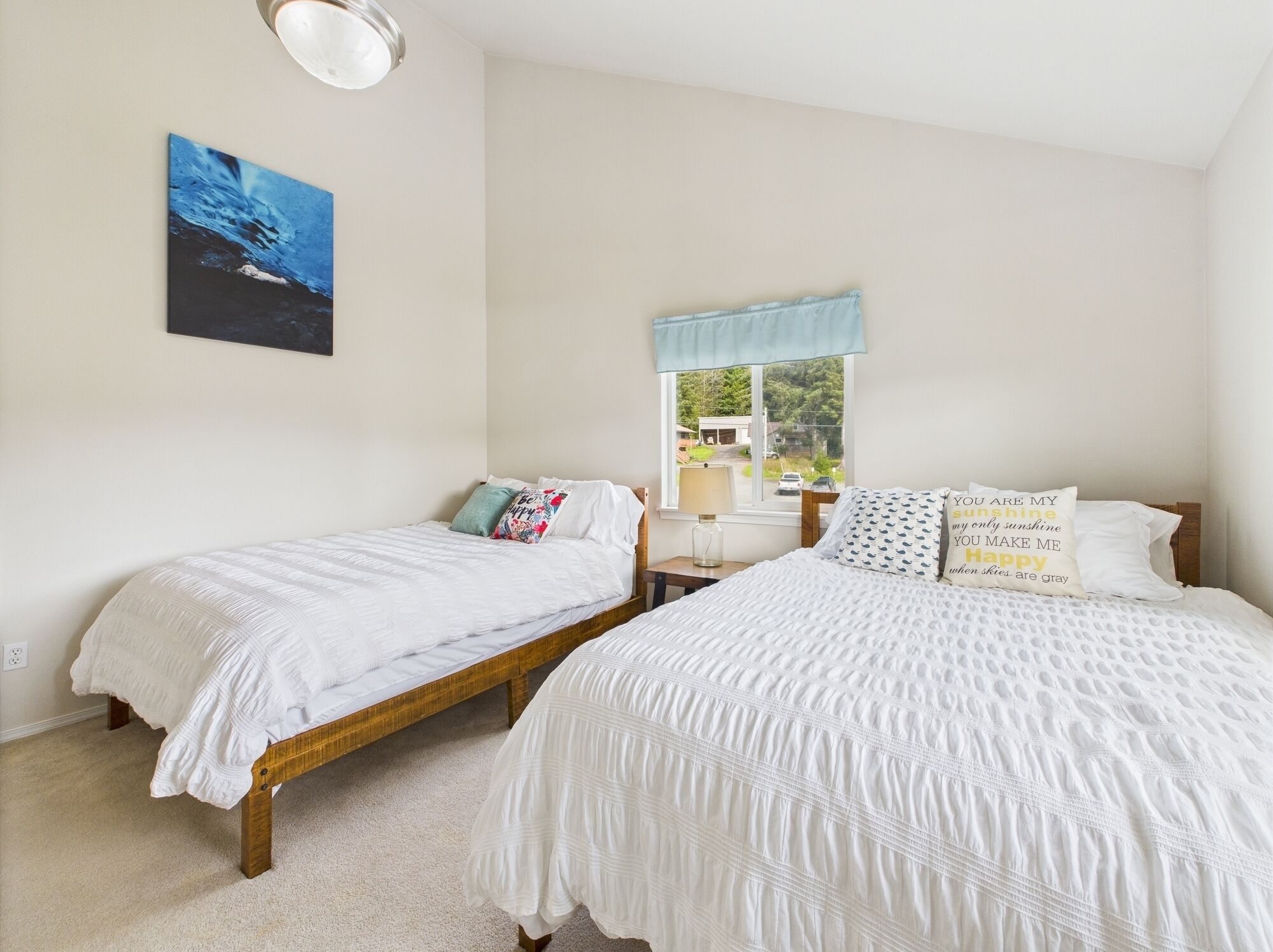 A bedroom with two single beds, a window with a view, a painting on the wall, and a bedside table with a lamp.