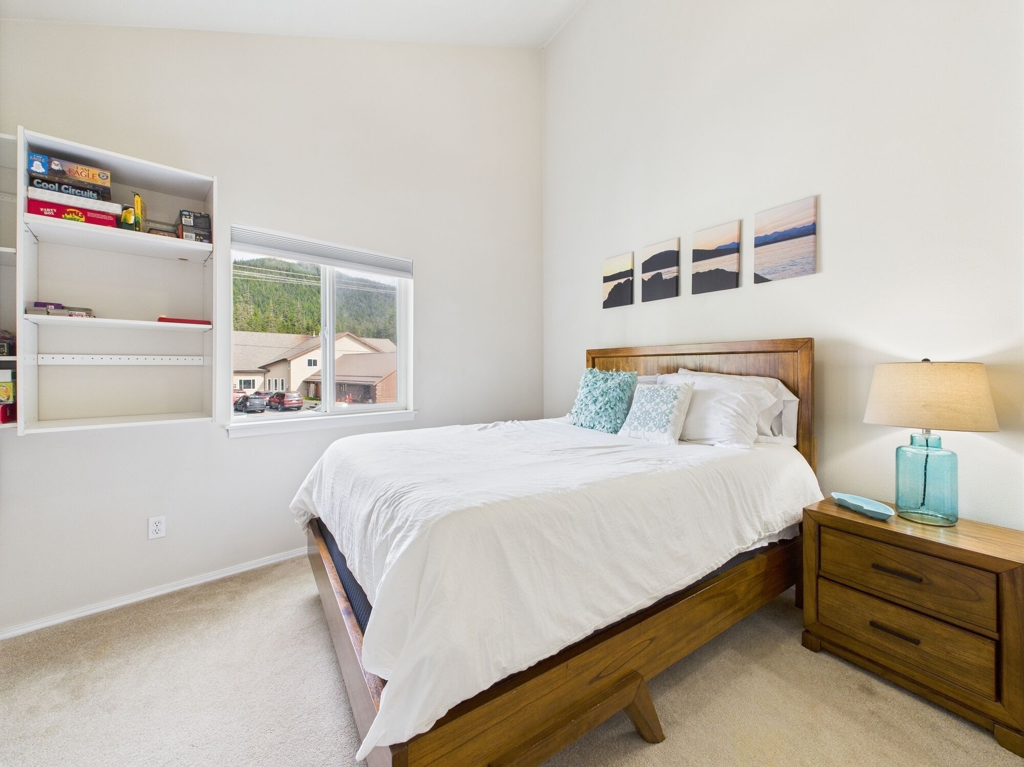 A bedroom with a wooden bed, white bedding, a nightstand with a lamp, and a shelf with books and a radio.