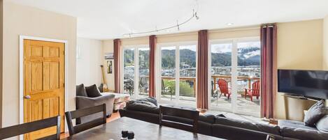 A spacious living room with a large dining table, comfortable seating, a TV, and a view of a marina and mountains through large windows.