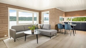 A modern living room with a grey sofa, a round coffee table, and a dining area with a table set for four. Large windows offer a view of a lake and hills. - Awesome home in Gjemnes with WiFi (Gjemnes)