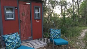 Una cabaña roja pequeña con un muelle de madera y dos sillas azules con cojines estampados.