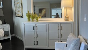 A living room with a white sofa, a white cabinet with drawers, a mirror above the cabinet, and a lamp on the right side. - Eloquently decorated 1 bedroom, 1 1/2  bath condo. Beautiful water views/balcony (West Palm Beach)