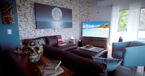 Una sala de estar con una pared de piedra, un televisor de gran tamaño que muestra una escena de playa, un sofá, una mesa de centro con libros y una calavera decorativa.