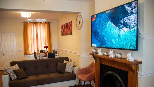 Een woonkamer met een bank, een open haard, een televisie en een eethoek op de achtergrond.
