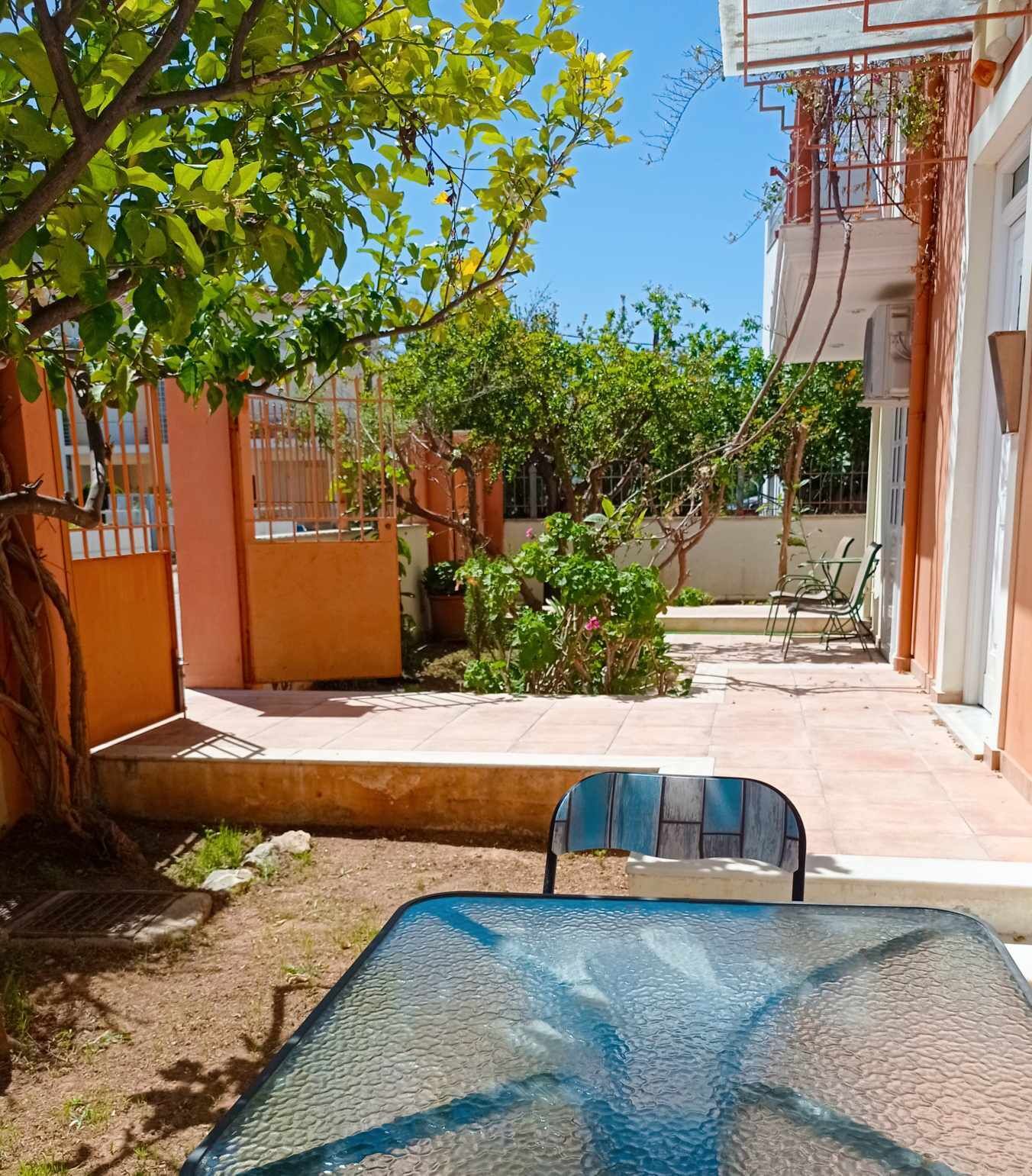 Une cour avec une table et des chaises en verre, entourée d’un bâtiment doté d’un portail en métal et d’un arbre.