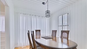 Une salle à manger avec une table ronde en bois, six chaises, des rideaux blancs et un luminaire suspendu.