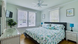 Une chambre à coucher comprenant un lit, deux tables de chevet, un ventilateur de plafond et une fenêtre avec des stores.