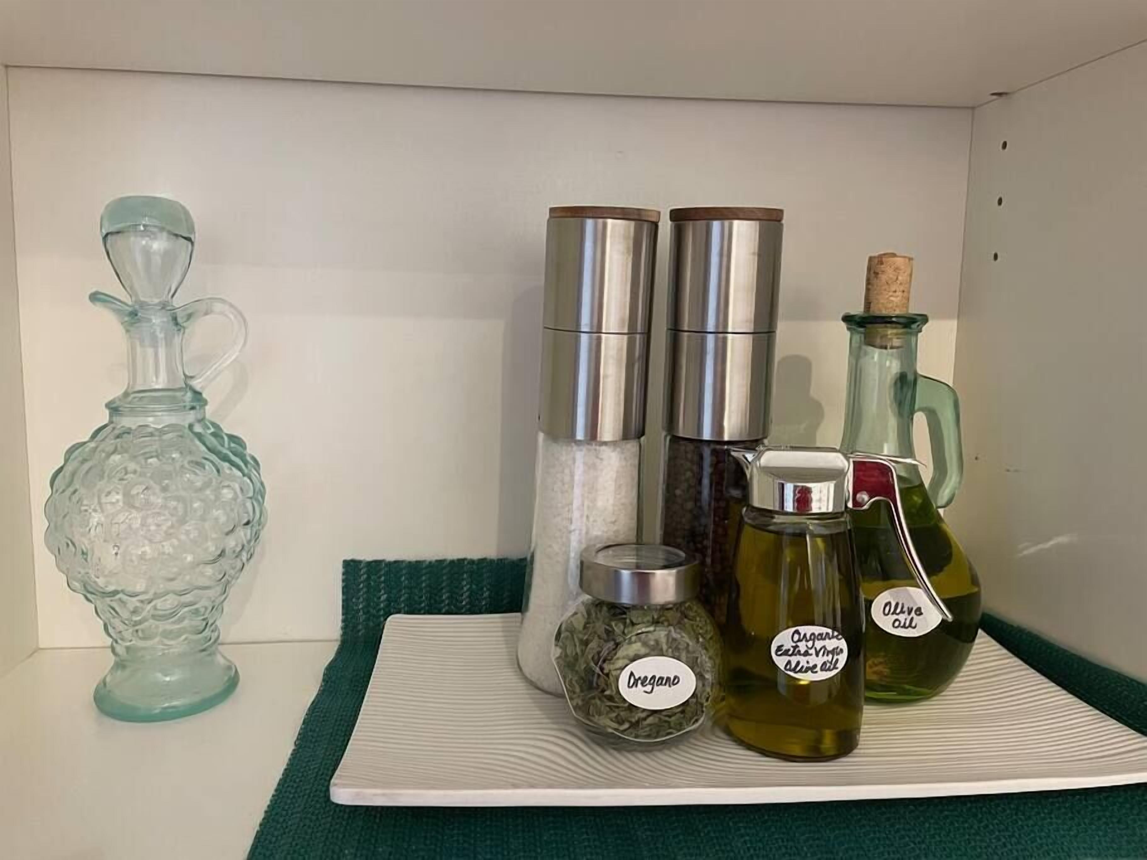 A shelf with various kitchen items including a glass pitcher, two stainless steel containers, a glass bottle with a cork, and a jar labeled "Dregano".