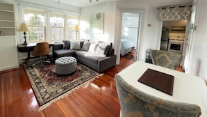 A spacious living room with a grey sectional sofa, a round ottoman, and a patterned rug. There is a dining area with a table and chairs, and a kitchen visible through an open doorway.
