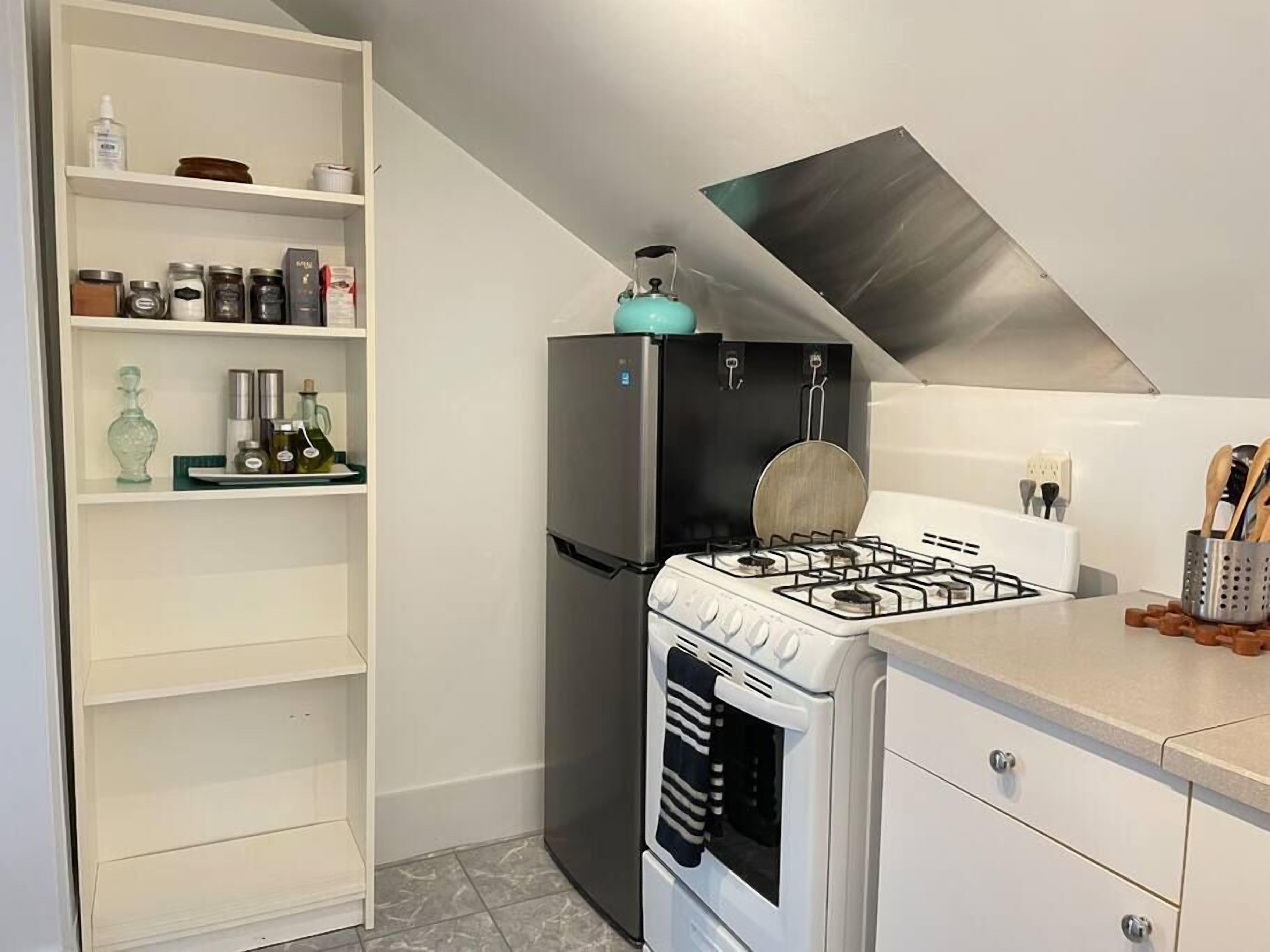 Una cocina compacta con un refrigerador blanco, una estufa a gas y un horno blanco. Hay repisas con utensilios de cocina y una pequeña encimera con utensilios.