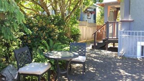 Un espace patio avec une table ronde et deux chaises, entouré d’un feuillage dense et d’une clôture en bois.
