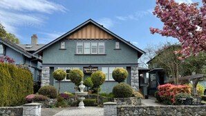 Un bâtiment à deux étages avec une façade en pierre, une enseigne indiquant « Trout Manor Touris Suites », et un jardin bien entretenu agrémenté d’une fontaine et d’arbustes taillés.
