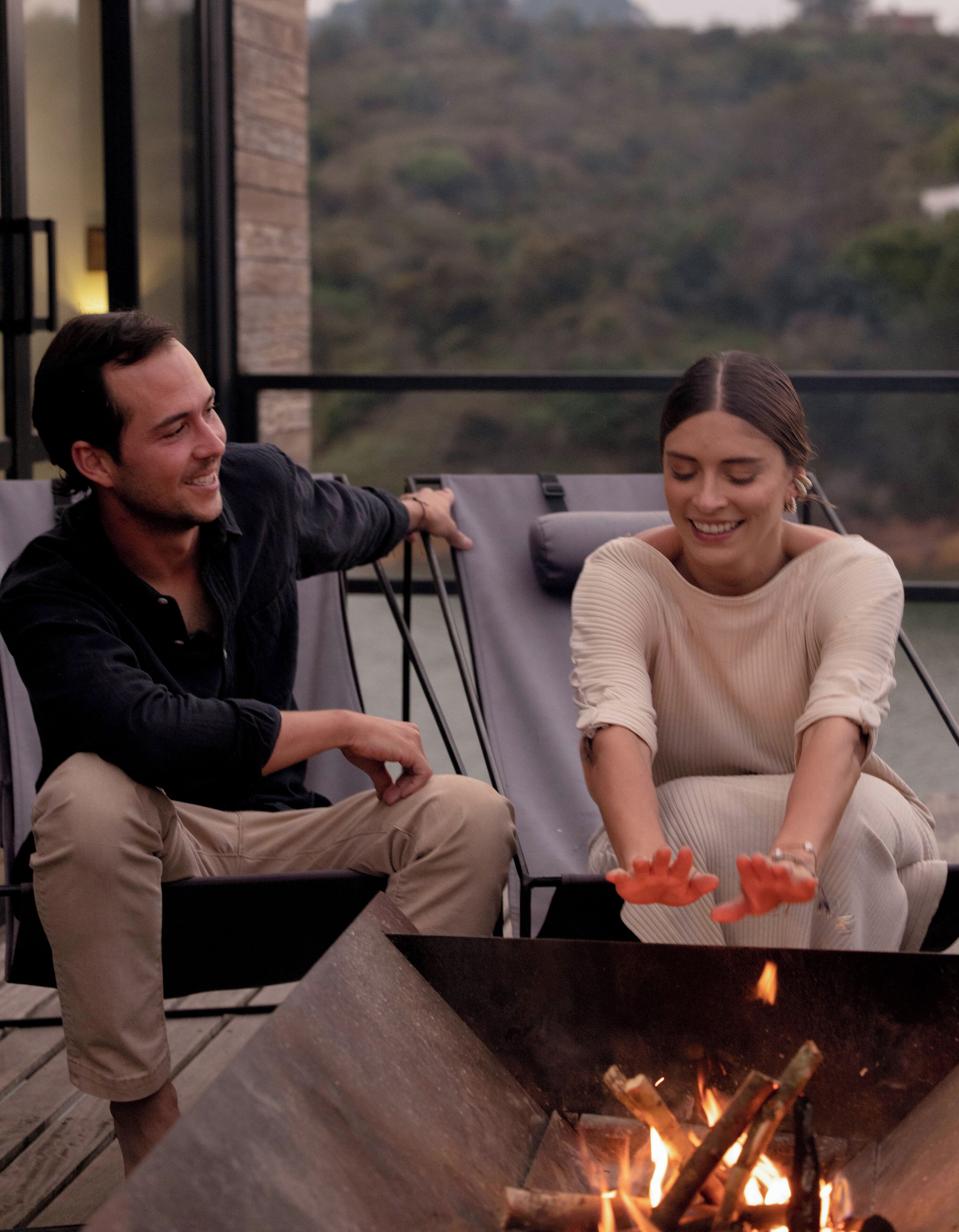 Dos personas sentadas junto a una fogata en un balcón con vistas a un paisaje montañoso.