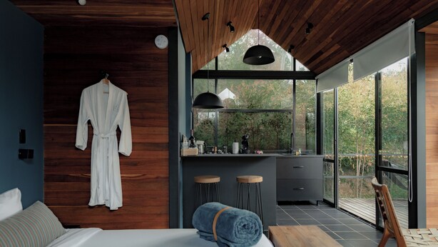 A modern bedroom with a large bed, a wooden ceiling, a robe hanging on the wall, and a view of the outdoors through large windows. - Boato Hotel (El Penol)