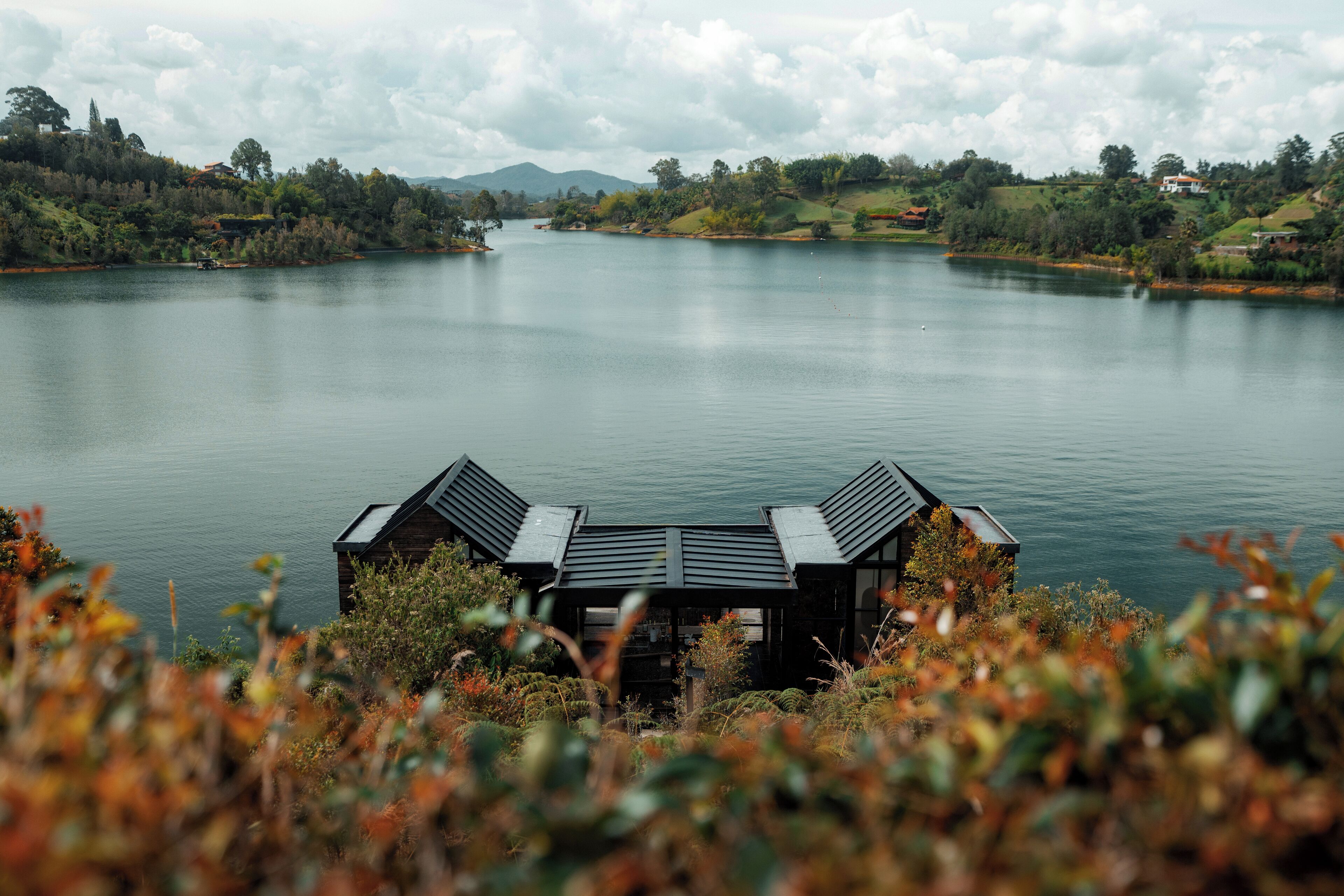 A lakeside cabin with a roof and a small deck, surrounded by lush greenery and a calm lake.