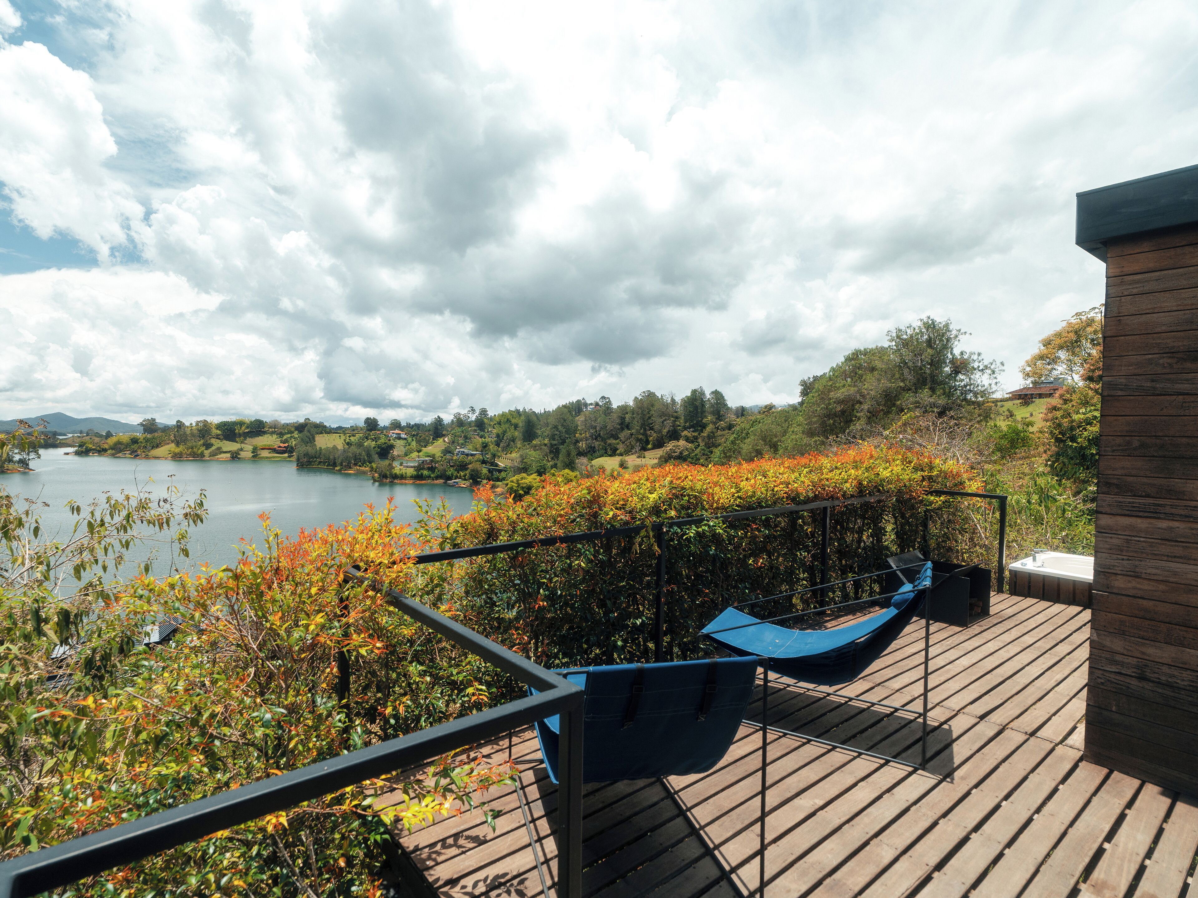 Terraza de madera con hamaca y bañera de hidromasaje con vistas a un lago y un paisaje boscoso al fondo.