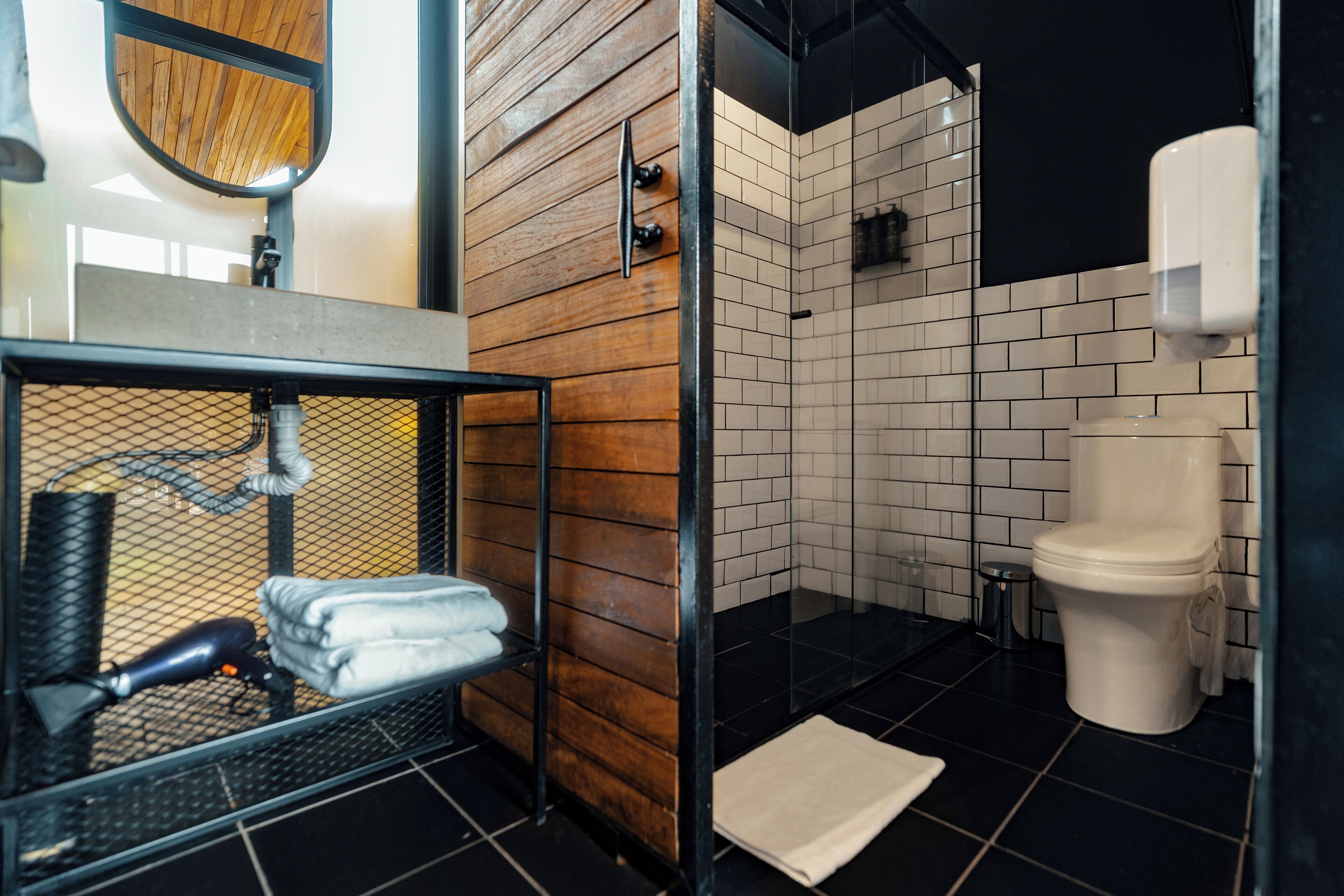 A modern bathroom with a wooden wall, a glass shower enclosure, a white toilet, and a metal sink with towels.