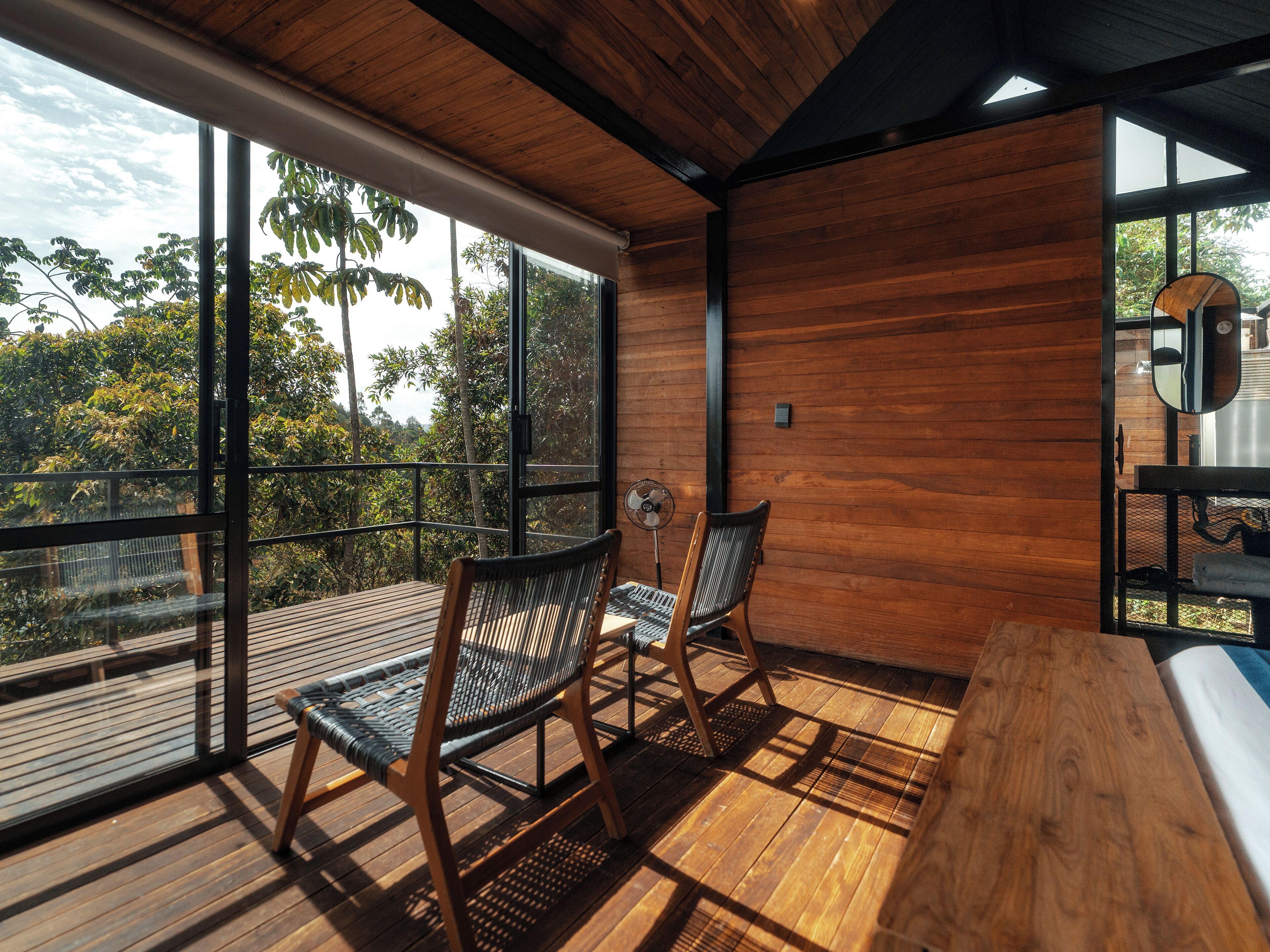 A balcony with wooden flooring and chairs, large windows with a view of trees, and a wooden wall.