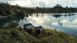 A lakeside cabin with a metal roof, surrounded by dense forest and hills in the background. - Boato Hotel (El Penol)