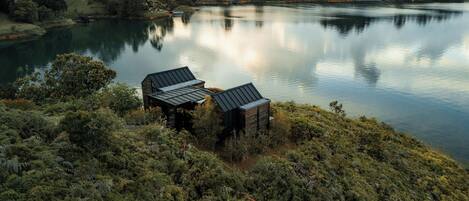 Una cabaña junto al lago, con techo de metal, rodeada de un bosque denso y colinas al fondo.