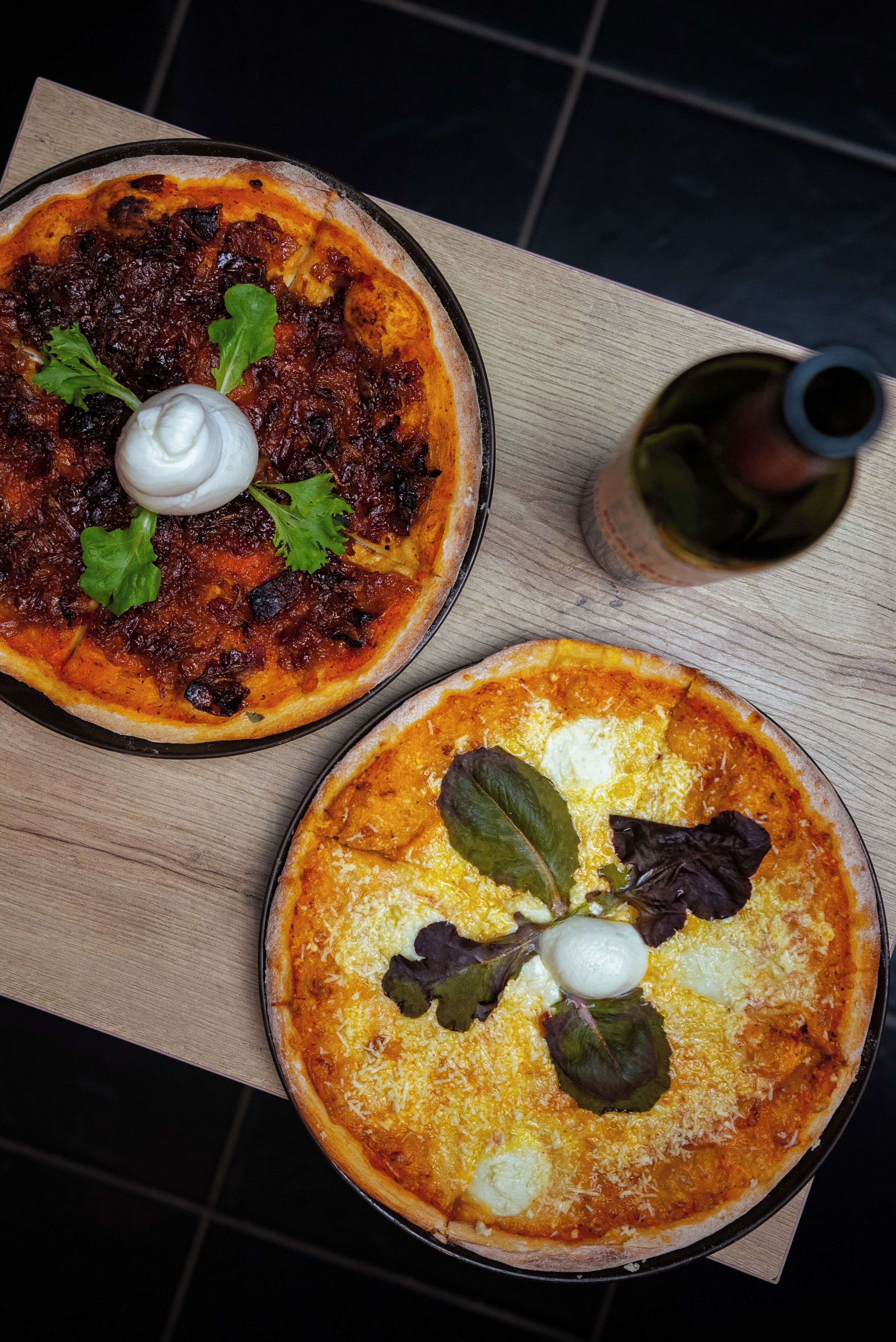 Two pizzas on a wooden table, one topped with a dark sauce and the other with cheese and greens.