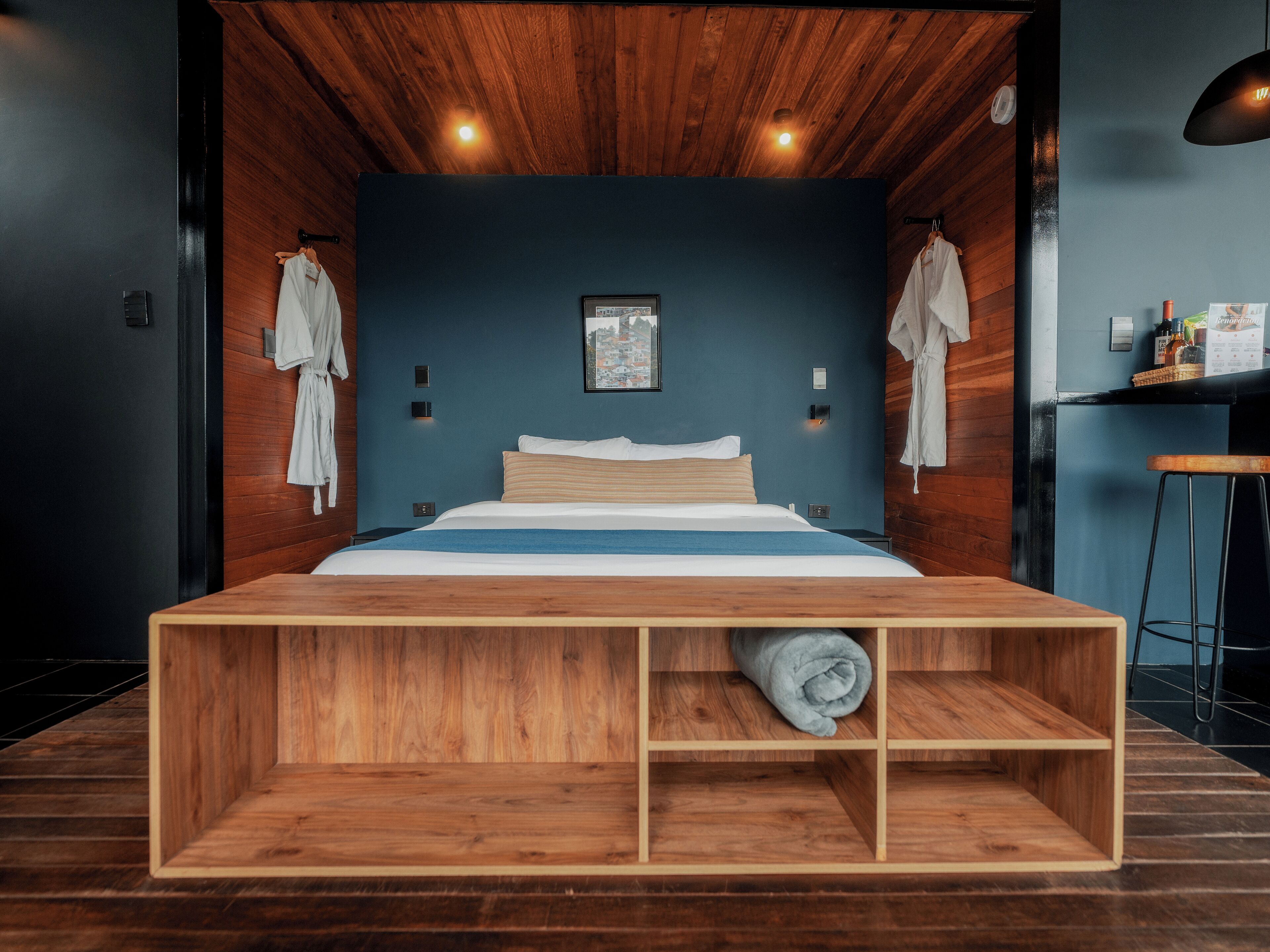 A modern bedroom with a wooden headboard, a bed with white and blue bedding, and a small wooden shelf with a rolled-up towel.